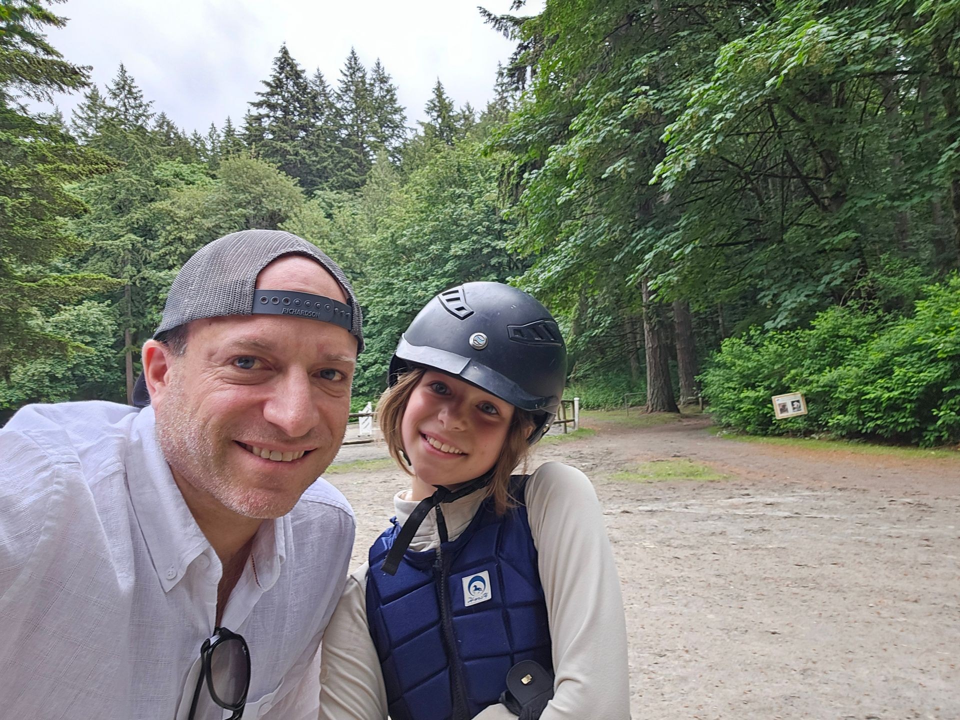 Smiling man and girl in equestrian helmet posing in wooded outdoor area.