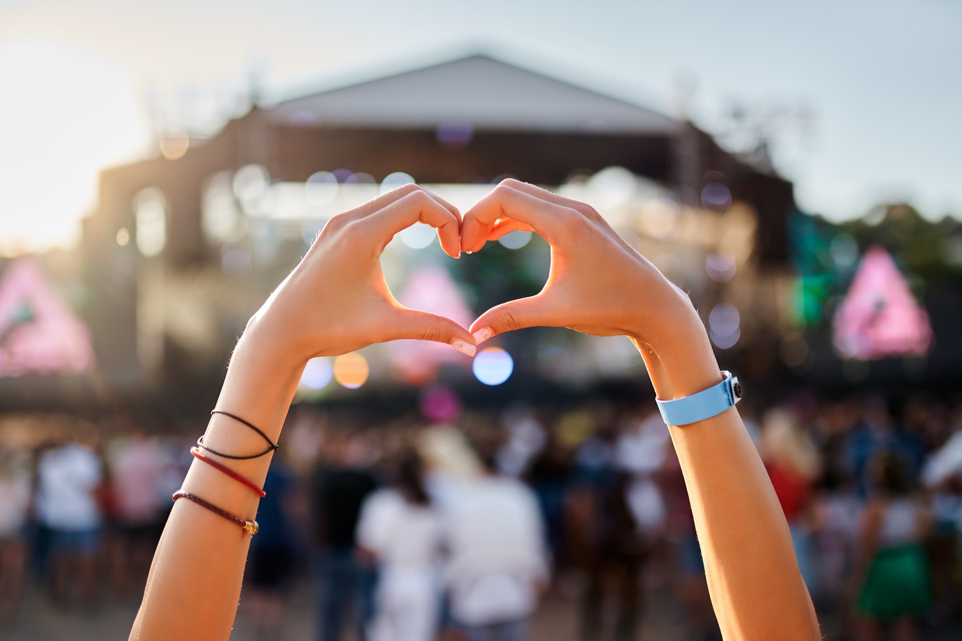 Hands shape heart sign at sunset beach music fest, crowd enjoys live concert. Outdoor summer event, happy fans party, love symbol, festive vibe by the sea. Silhouette, sun glow, entertainment.